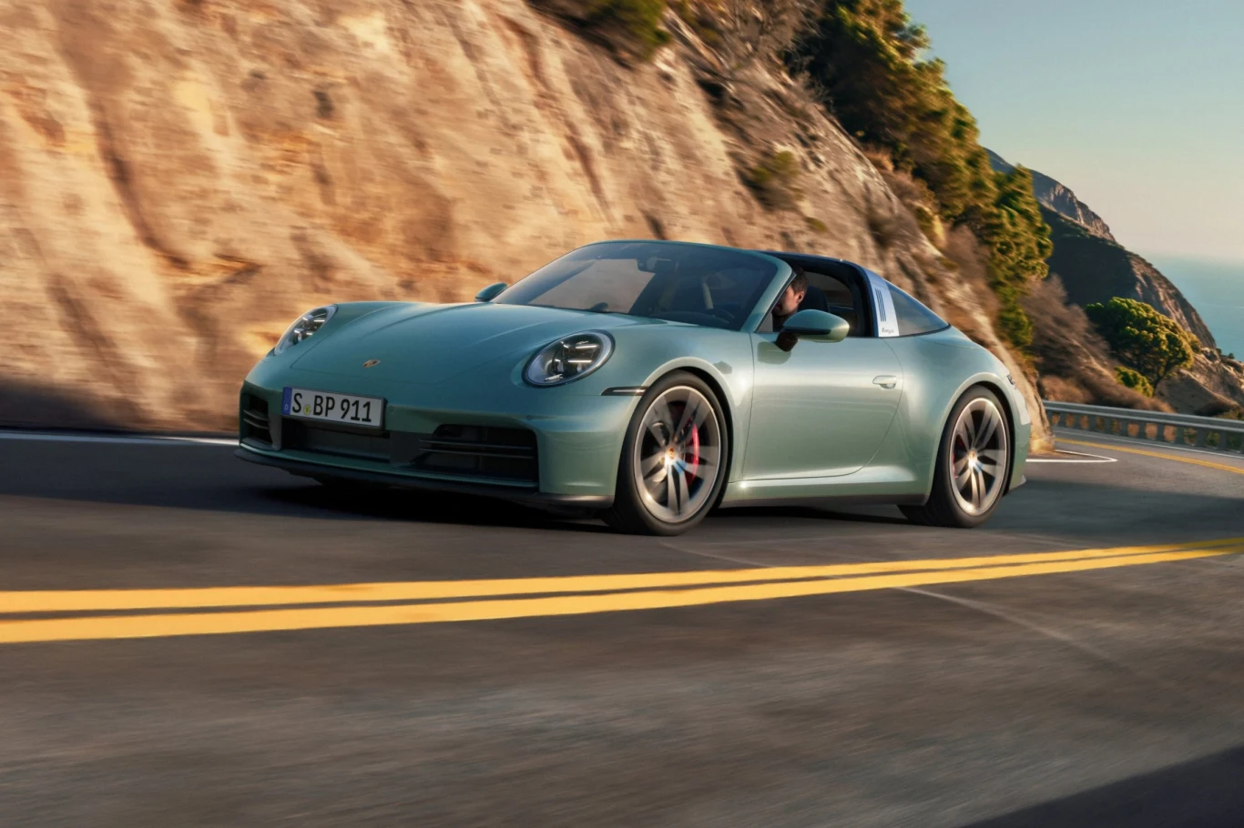Porsche 911 Targa 4S ma teraz 480 KM Jasnozielony sportowy samochód marki Porsche jedzie dynamicznie krętą asfaltową drogą wśród skalistego górskiego krajobrazu, przy zachodzącym słońcu, po żółtych liniach drogowych, na tle nadmorskich klifów i zieleni.