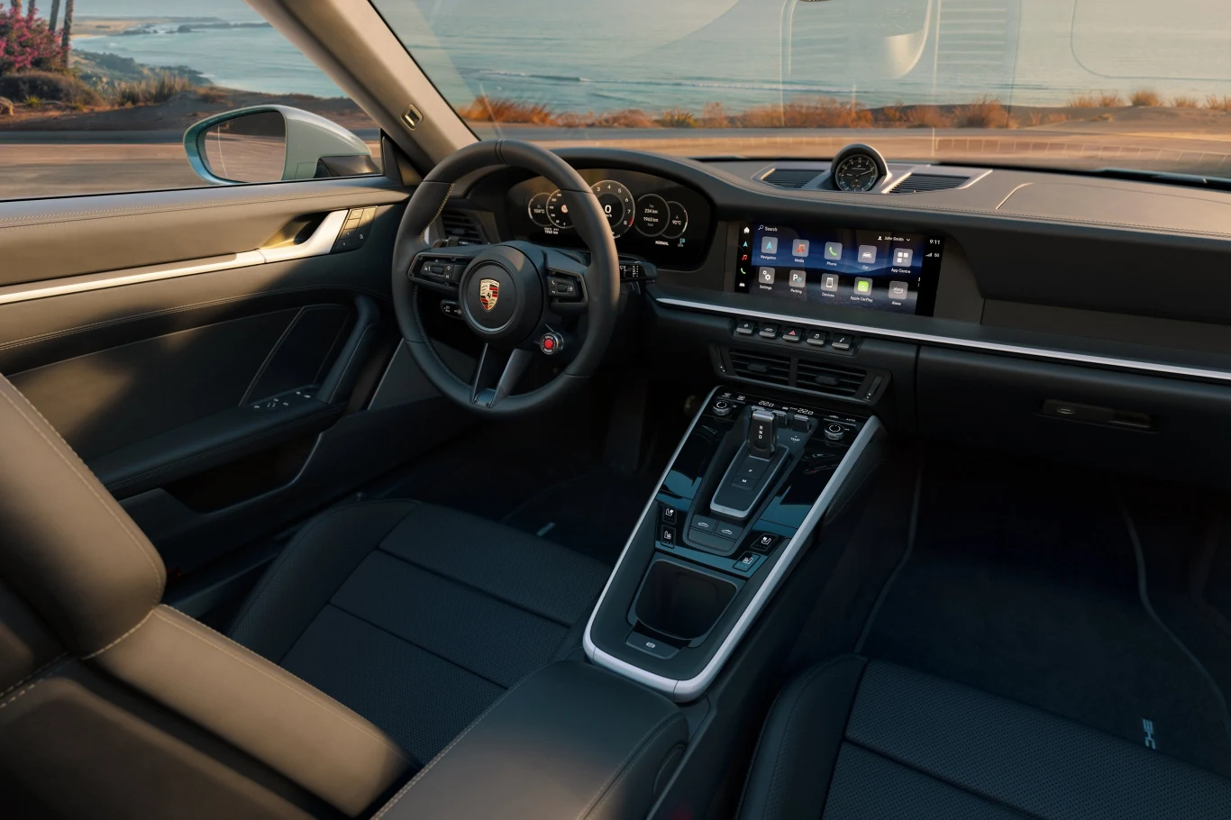 Porsche 911 Carrera 4S Wnętrze luksusowego samochodu sportowego z dużym dotykowym ekranem multimedialnym, sportową kierownicą i skórzanymi siedzeniami, widok przez przednią szybę na drogę oraz wybrzeże.