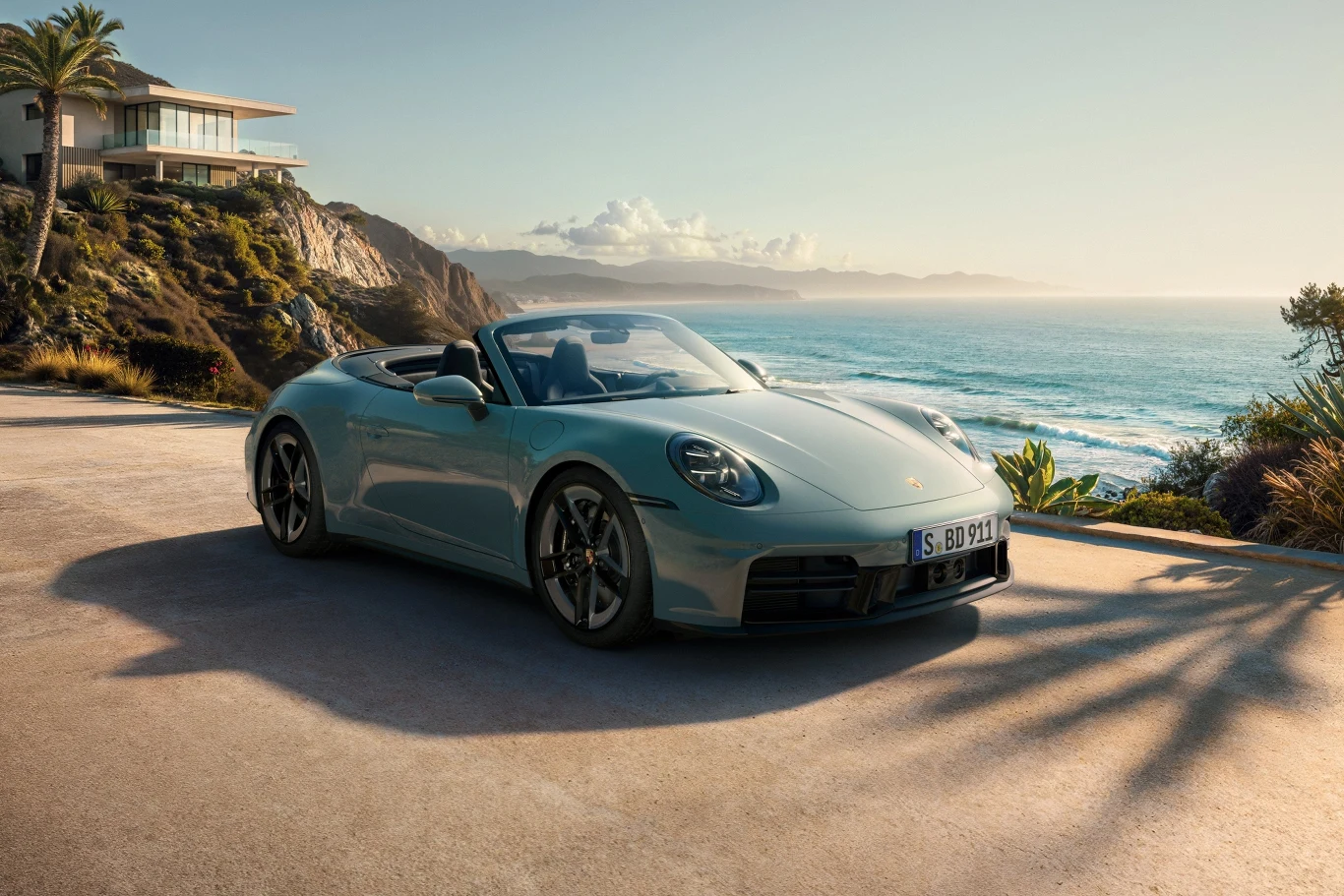 Porsche 911 Carrera 4S Cabriolet Srebrny kabriolet Porsche zaparkowany przy nadmorskiej drodze z widokiem na klify i ocean, w tle nowoczesna willa na wzgórzu oraz bujna roślinność i palmy podkreślające luksusowe otoczenie.