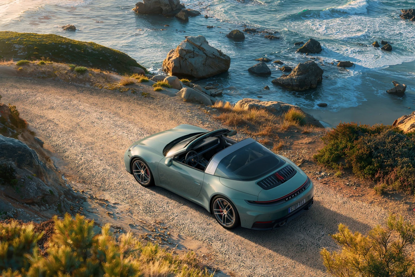 Porsche 911 Targa 4S Luksusowy kabriolet sportowy porusza się żwirową drogą nad skalistym wybrzeżem oceanu, w tle rozbijające się o brzegi fale i słoneczny krajobraz z roślinnością śródziemnomorską.