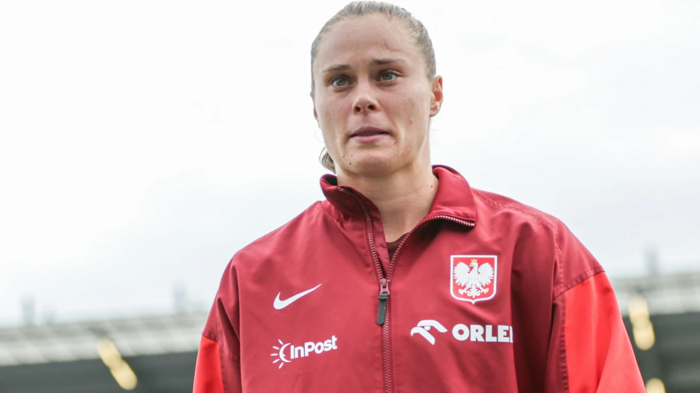 Kobieta w czerwonej kurtce sportowej z godłem Polski i logotypami sponsorów stoi na tle zamazanego stadionu, patrząc przed siebie.