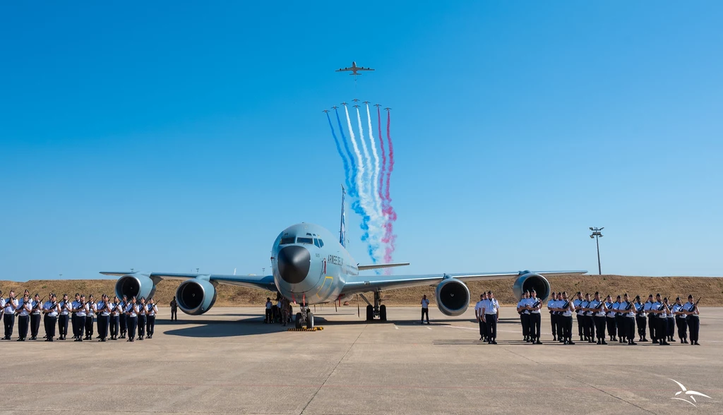 Pożegnalny przelot tankowca KC-135 w Siłach Powietrznych Francji Wojskowy samolot transportowy stoi na płycie lotniska, po obu stronach zgromadzeni są członkowie orkiestry wojskowej w galowych mundurach, a nad samolotem przelatuje grupa samolotów akrobacyjnych zostawiających barwny ślad dymu na niebie.