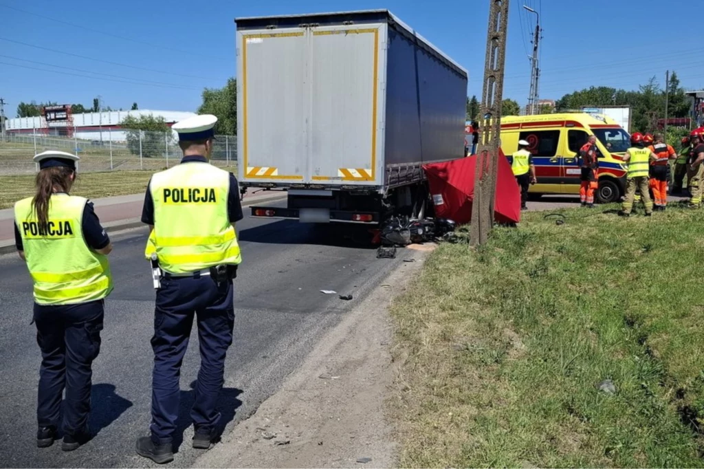 Drogowy wypadek z udziałem ciężarówki i samochodu osobowego, obecność policjantów w kamizelkach odblaskowych oraz ekipy ratunkowej udzielającej pomocy, na poboczu rozmieszczone służby ratunkowe i ambulans.