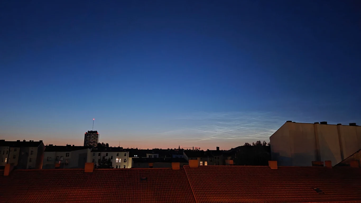 Obłoki srebrzyste widoczne po zmierzchu Panorama miasta z widokiem na dachy budynków i wieżowiec z sygnalizacją świetlną przeciwlotniczą, na tle ciemniejącego, niebieskiego nieba o zmierzchu.