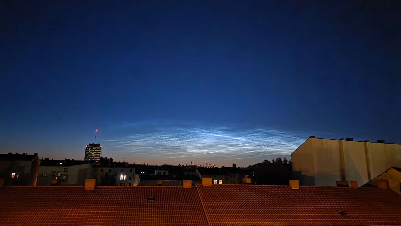 Im ciemniej, tym obłoki srebrzyste były lepiej widoczne Nocne niebo z delikatnie podświetlonymi chmurami nad miejską panoramą, na pierwszym planie dachy budynków, z lewej strony wyróżnia się wieżowiec z czerwonym światłem na szczycie.