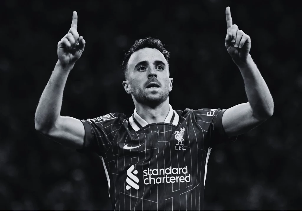 Diogo Jota Piłkarz w koszulce Liverpoolu w dynamicznej pozie unosi obie ręce z wyprostowanymi palcami wskazującymi w górę, tło rozmyte, całość w odcieniach czerni i bieli.