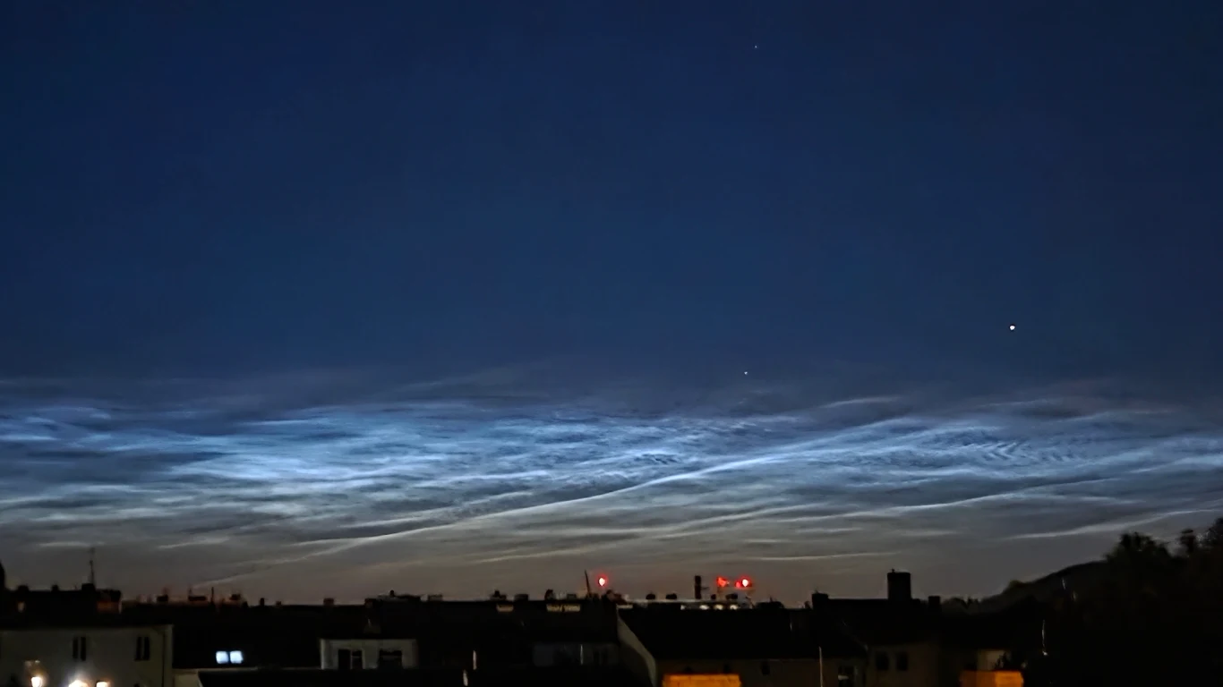 Obłoki srebrzyste widoczne 2 lipca w zachodniej Polsce Nocne niebo z widocznymi obłokami srebrzystymi nad ciemną sylwetką zabudowań miejskich, kilka świateł na dachach budynków po lewej stronie, delikatne fale chmur prześwitujące w błękicie nieba