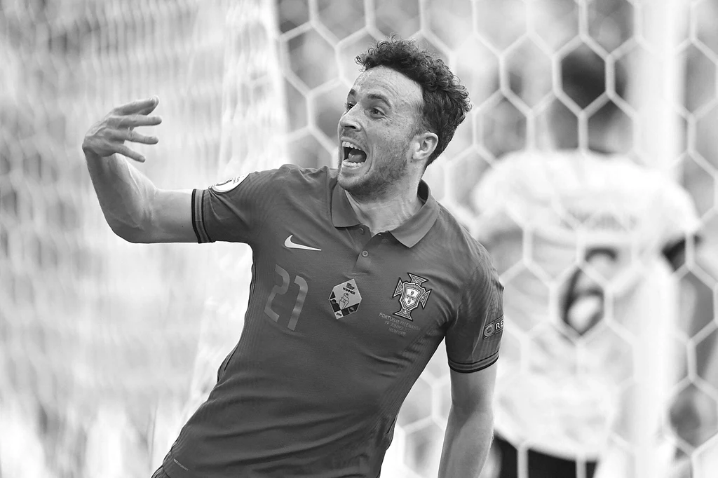 Diogo Jota Piłkarz w koszulce z emblematem Portugalii świętuje zdobycie gola na tle siatki bramki, wyrażając emocje szerokim gestem ręki i żywą mimiką twarzy.