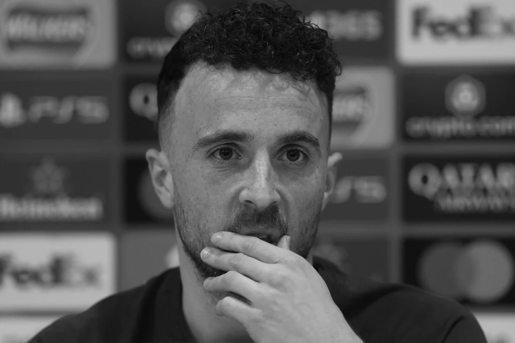 Diogo Jota Mężczyzna o kręconych włosach z zarostem, opierający rękę na ustach, w tle rozmyte logotypy sponsorów sportowych.