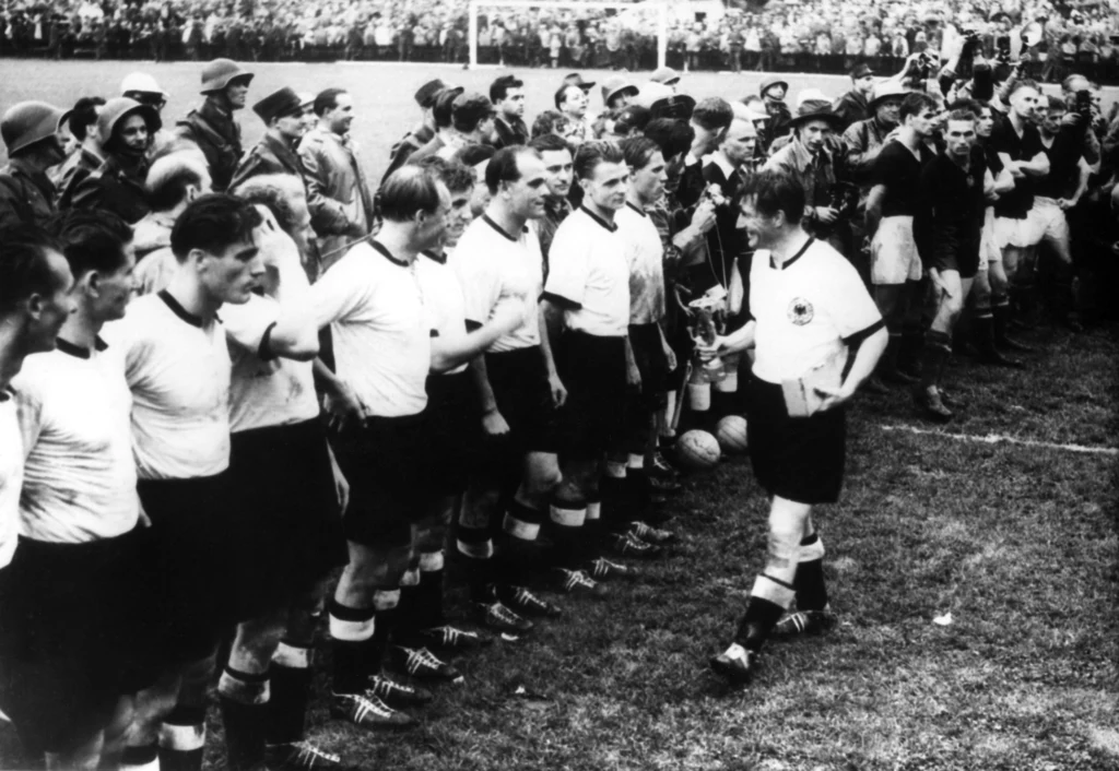 Fritz Walter i reszta reprezentacji Niemiec świętująca zdobycie MŚ 1954 Piłkarze ustawieni w szeregu na boisku podczas ceremonii lub prezentacji drużyny, wokół nich zgromadzeni widzowie i oficjele, atmosfera sportowego wydarzenia z historycznego okresu.