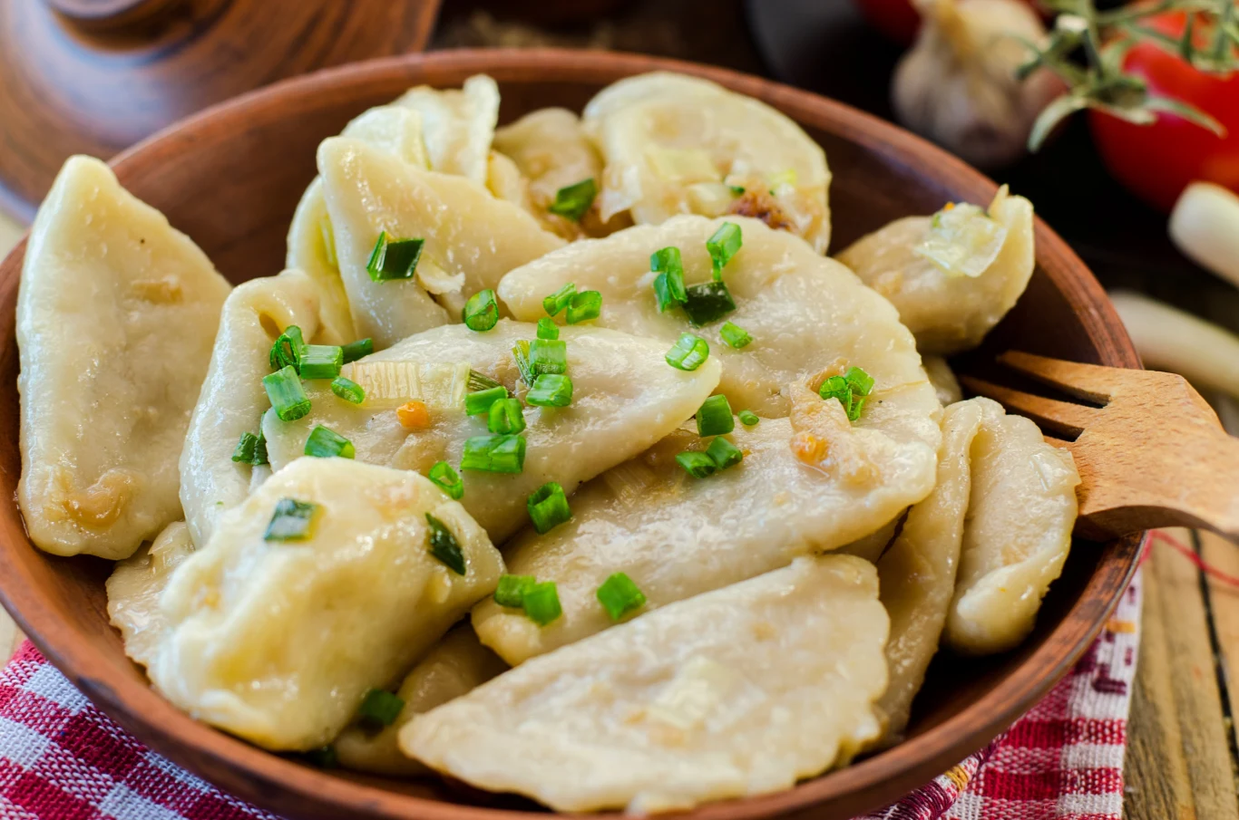 Pierogi z bobem. Poznaj przepis od Ewy Wachowicz Pierogi podane w glinianej misce, udekorowane świeżo posiekanym szczypiorkiem; po prawej stronie drewniany widelec, w tle wyraźnie zaznacza się kuchenny klimat.