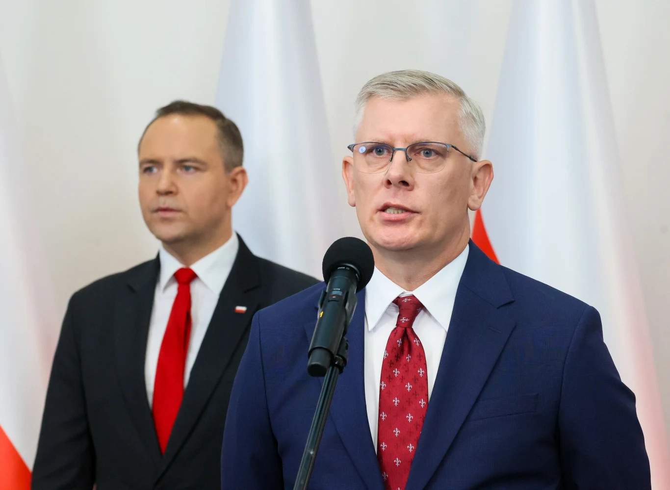 Sławomir Cenckiewicz (po prawej) został zapowiedziany przez Karola Nawrockiego jako przyszły szef BBN Dwóch mężczyzn w garniturach stoi na tle biało-czerwonych flag, jeden przemawia do mikrofonu, drugi stoi w tle.