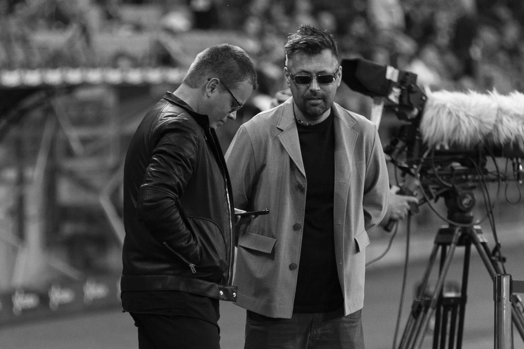 Jarosław Królewski, Thomas Kokosinski Dwóch mężczyzn w rozmowie na tle profesjonalnej kamery i rozmytego stadionu, jeden z nich skupiony na telefonie, drugi patrzy przed siebie, obaj w casualowych ubraniach.