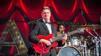 Muzyk w eleganckim garniturze grający na czerwonej gitarze elektrycznej na tle podświetlonej sceny, w tle perkusista oraz elementy scenografii z czerwonymi zasłonami.