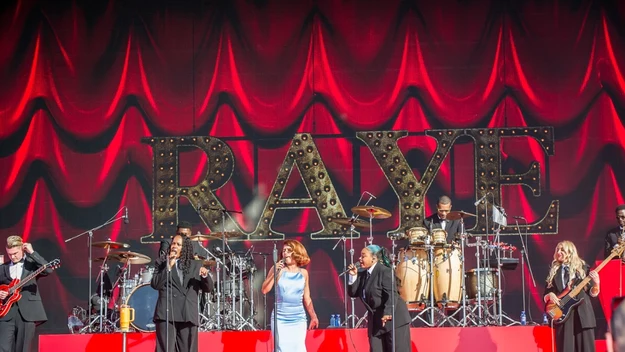 Grupa muzyków wykonuje koncert na scenie z instrumentami przy dużym napisie RAYE na tle czerwonej kotary, na pierwszym planie wokalistka w jasnej sukni i śpiewający z nią artyści.