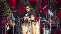 Dwóch muzyków na scenie, jeden gra na zestawie instrumentów perkusyjnych, a drugi śpiewa do mikrofonu, oboje ubrani elegancko w garnitury, w tle duży, kolorowy ekran.
