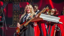Uśmiechnięta kobieta grająca na gitarze basowej na scenie, w tle widoczny klawiszowiec oraz muzycy grający na instrumentach dętych, scena ozdobiona czerwonymi dekoracjami scenograficznymi.
