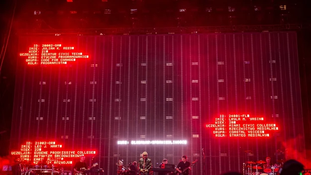Scena koncertowa z zespołem muzycznym występującym na tle dużego ekranu, na którym w czerwonym kolorze wyświetlają się dane tekstowe; widoczne instrumenty, światła i skupiona na wydarzeniu publiczność.