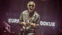 Mężczyzna w okularach przeciwsłonecznych stoi na scenie, trzymając mikrofon, ubrany w czarną, błyszczącą kurtkę i naszyjnik z symbolem ankh, za nim kolorowe światła oraz rozmyte litery na ekranie.