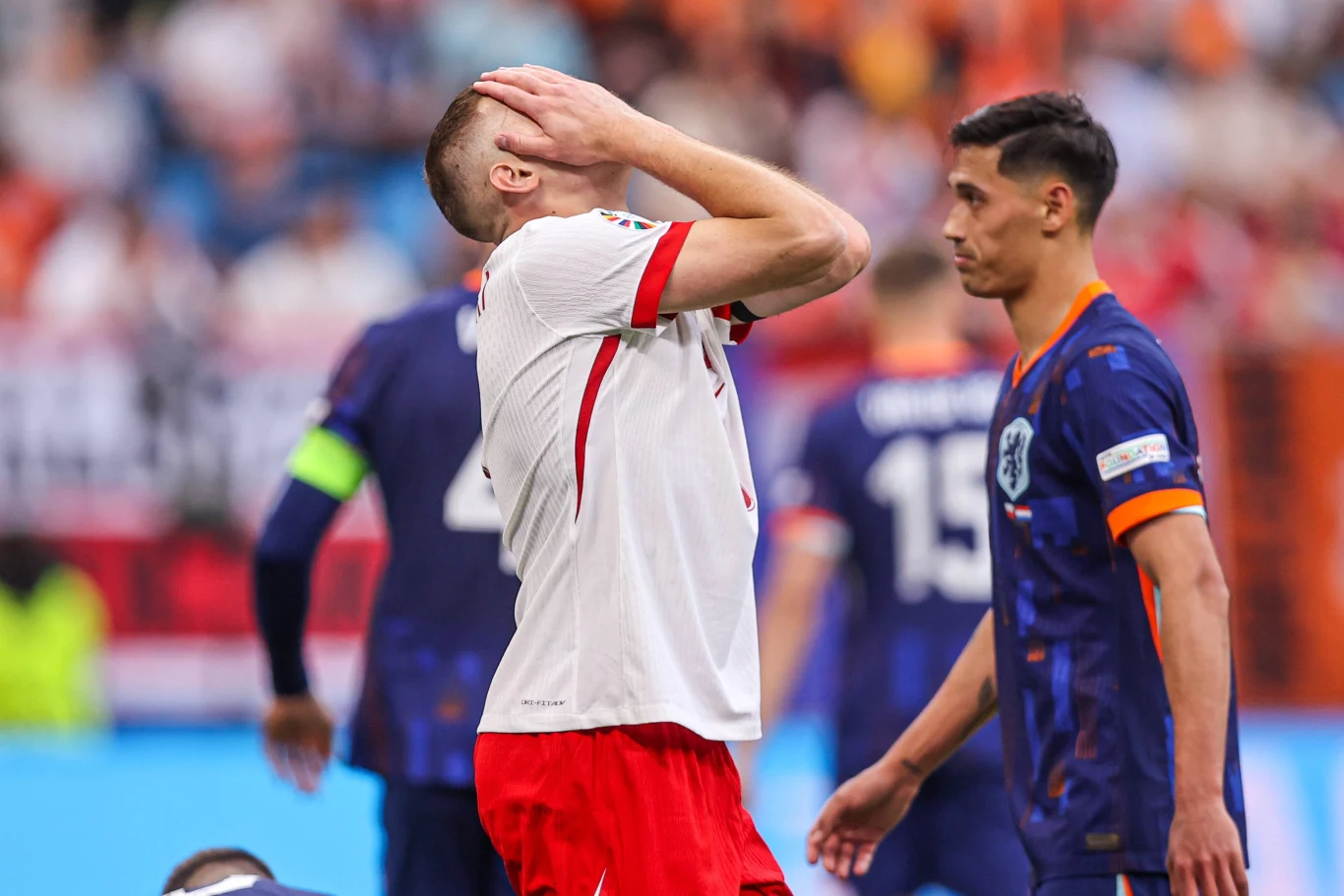 Piłkarz w białej koszulce i czerwonych spodenkach trzyma głowę w geście rozczarowania, w tle zawodnicy drużyny przeciwnej w granatowych strojach oraz rozmyty tłum na trybunach.