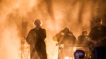 Grupa muzyków wykonuje koncert na scenie w mocnym świetle i gęstej mgle, centralną postacią jest śpiewający wokalista, wokół znajdują się instrumenty i sprzęt muzyczny.