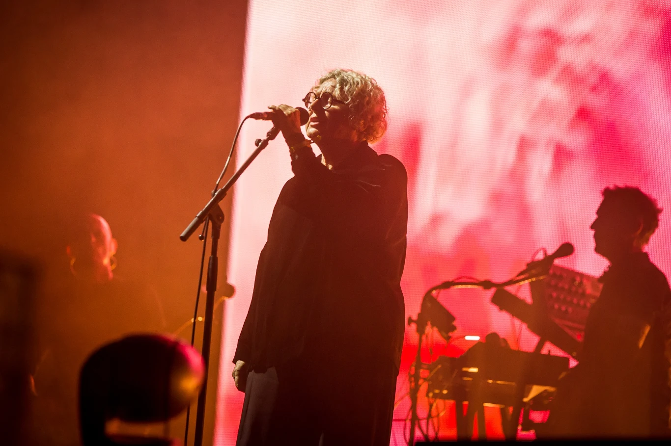 Wokalista z mikrofonem stoi na scenie oświetlonej intensywnym, czerwonym światłem, w tle widoczni muzycy obsługujący elektroniczne instrumenty.