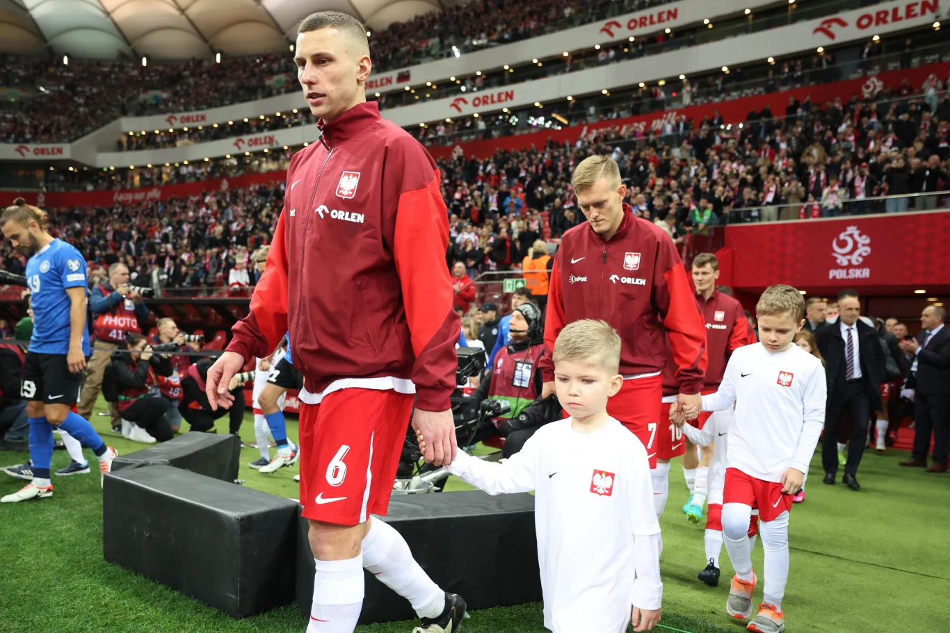 Piłkarze reprezentacji Polski w czerwonych dresach wychodzą na boisko prowadzając dzieci, w tle wypełnione trybuny stadionu i widoczne flagi biało-czerwone.