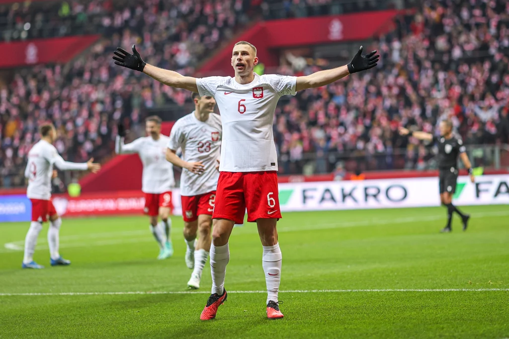 Jakub Piotrowski Piłkarz w biało-czerwonym stroju, z numerem 6 i rozłożonymi ramionami, świętuje na stadionie w otoczeniu innych zawodników oraz tłumu kibiców na trybunach.
