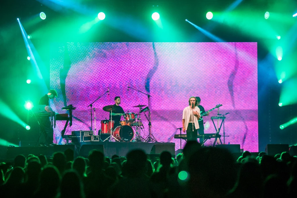 Wiktor Waligóra na Open'er Festivalu 2025 Grupa muzyków występuje na scenie koncertowej z dużym, kolorowym ekranem LED w tle, oświetlenie sceniczne w intensywnych odcieniach zieleni i fioletu, przed sceną liczna widownia.