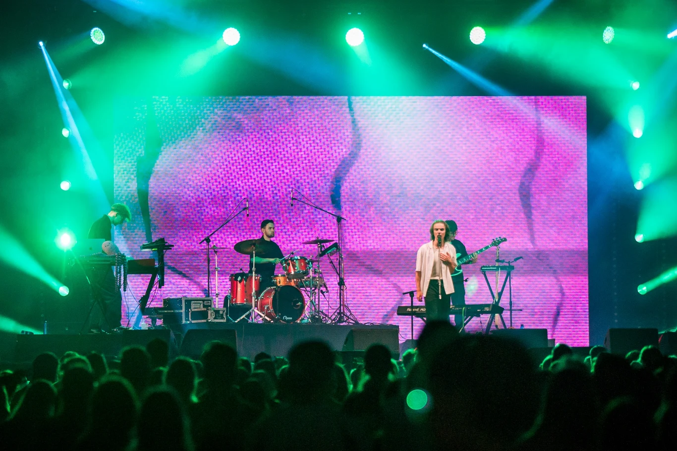 Grupa muzyków występuje na scenie koncertowej z dużym, kolorowym ekranem LED w tle, oświetlenie sceniczne w intensywnych odcieniach zieleni i fioletu, przed sceną liczna widownia.