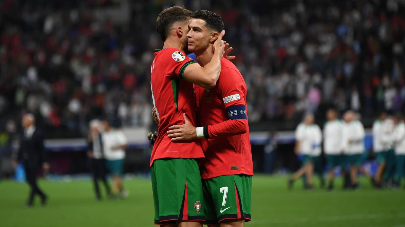 Cristiano Ronaldo żegna Diogo Jotę Dwóch piłkarzy w czerwono-zielonych strojach obejmuje się na środku boiska, w tle widoczna rozmyta publiczność oraz kilku innych zawodników.