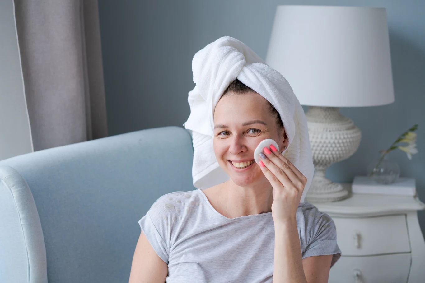 Uśmiechnięta kobieta nakłada kosmetyk na twarz za pomocą płatka kosmetycznego, włosy owinięte białym ręcznikiem, w tle jasna lampa i dekoracyjny kwiat na stoliku.