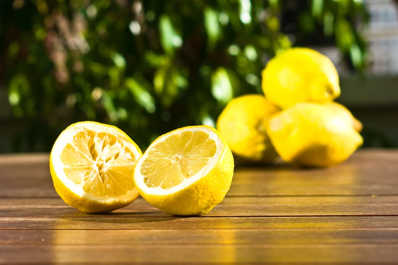 Połówka świeżo przeciętej cytryny oraz kilka całych cytryn na drewnianym stole z rozmytym zielonym tłem.