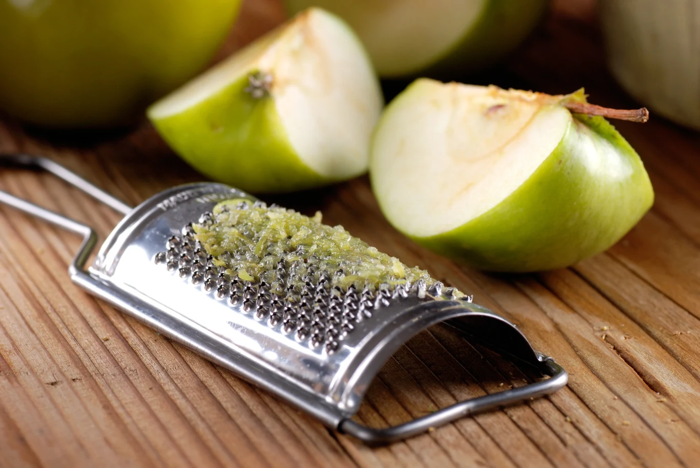 Zielone jabłko pokrojone na ćwiartki leży na drewnianym blacie obok metalowej tarki z utartym miąższem jabłka.