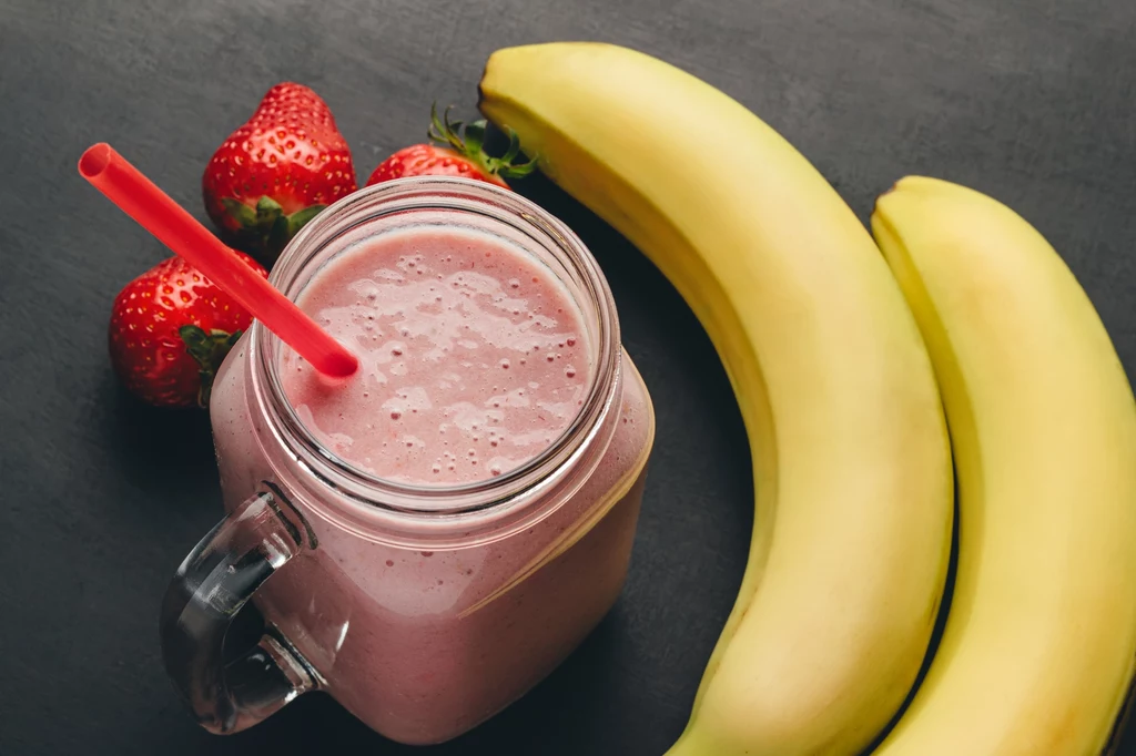 Przygotowujesz koktajl? Nie wyrzucaj skórek z banana, wykorzystaj je jako nawóz do róż! Koktajl owocowy w szklanym słoiku z czerwonym słomką otoczony dwoma bananami oraz kilkoma świeżymi truskawkami na ciemnym tle.