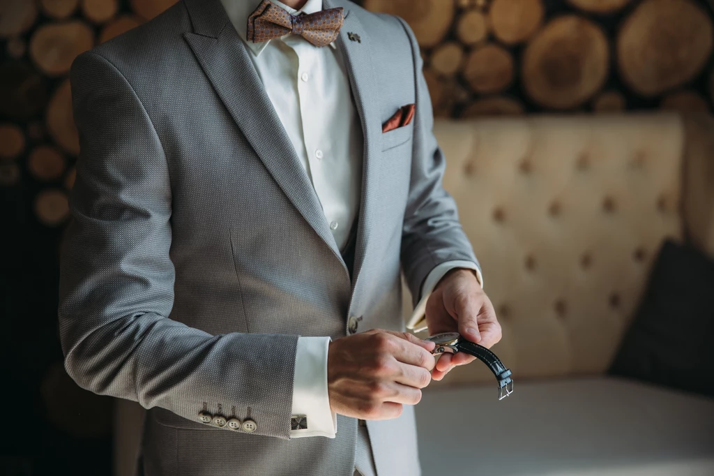 Męskie stylizacje na wesele Elegancko ubrany mężczyzna w szarym garniturze z muchą zakłada zegarek na rękę, tło stanowi jasna sofa oraz dekoracja z drewnianych okręgów.