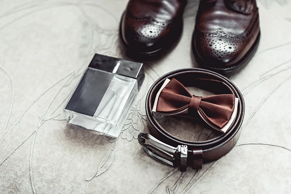 Dodatki do garnituru Skórzane eleganckie buty, flakon perfum, brązowa mucha oraz pasek na jasnym tle, stanowiące zestaw akcesoriów męskich przygotowanych do formalnej stylizacji.