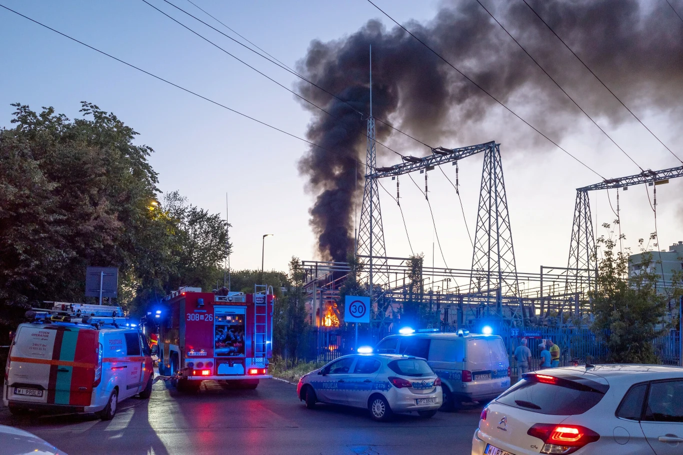 Pożar stacji transformatorowej na Pradze-Południe Straż pożarna i policja interweniują przy dużym pożarze na terenie stacji elektroenergetycznej, z której wydobywa się gęsty, czarny dym, wokół zgromadziło się kilka pojazdów służbowych z włączonymi sygnałami świetlnymi.