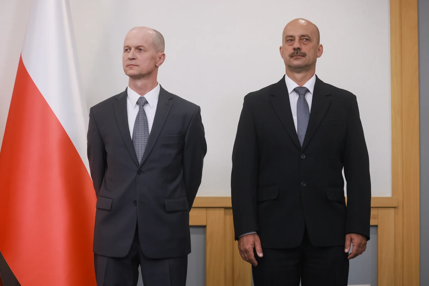 Desygnowani na zastępców szefa BBN gen. bryg. Mirosław Bryś (po prawej) i gen. bryg. Andrzej Kowalski (po lewej) Dwóch mężczyzn w garniturach stoi obok siebie na tle polskiej flagi i drewnianej ściany, obaj mają poważne wyrazy twarzy i patrzą w różnych kierunkach.