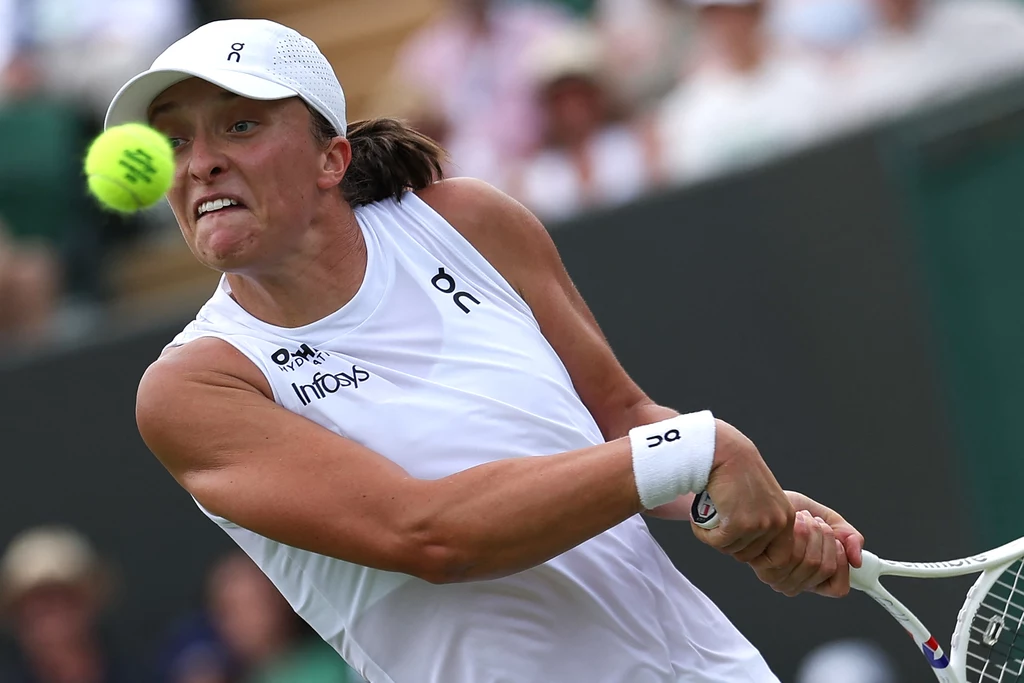 Iga Świątek w trakcie Wimbledonu Zawodniczka ubrana w biały strój sportowy oraz czapkę, skoncentrowana podczas odbicia piłki tenisowej rakietą, w tle rozmyta publiczność na trybunach.