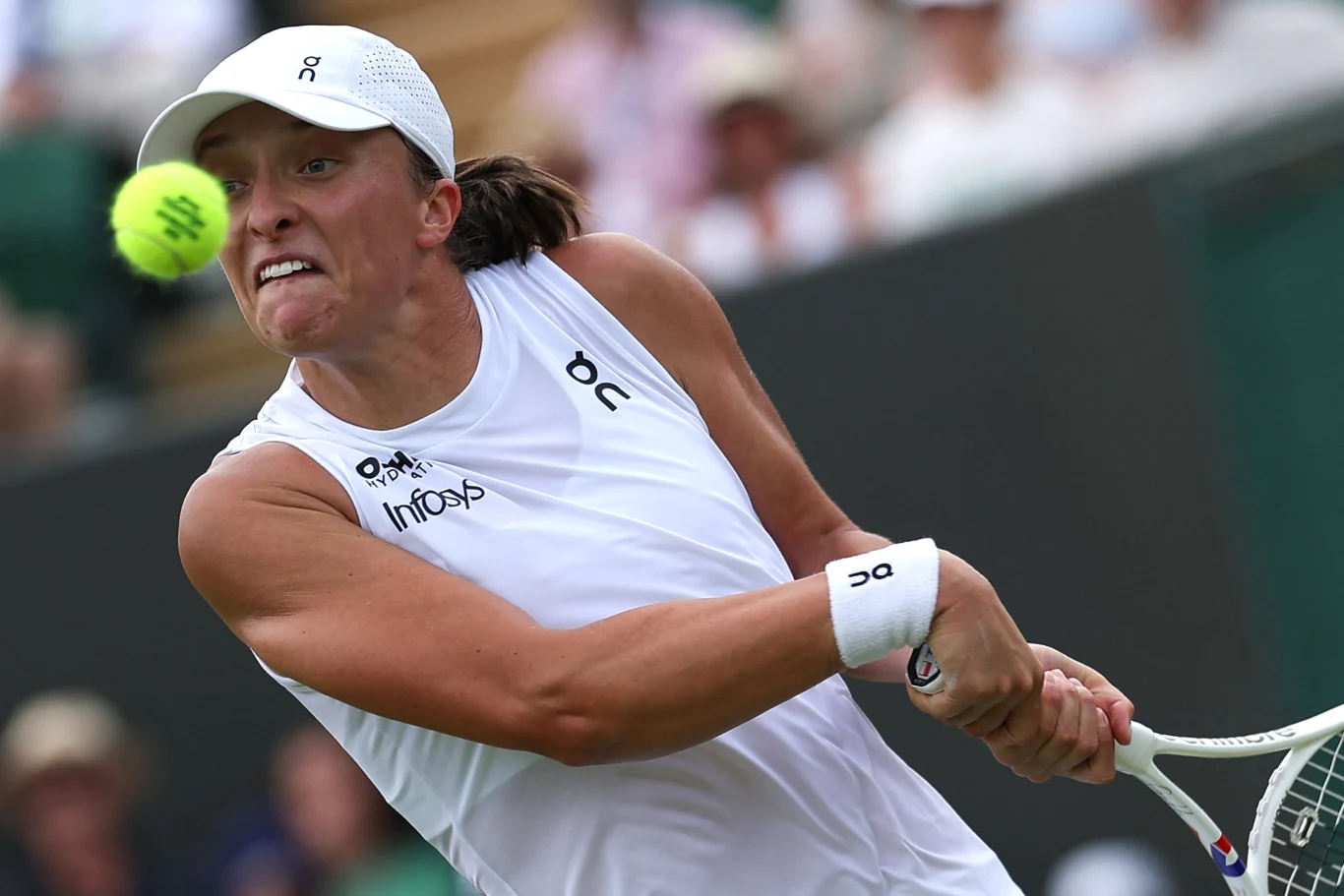 Iga Świątek w trakcie Wimbledonu Zawodniczka ubrana w biały strój sportowy oraz czapkę, skoncentrowana podczas odbicia piłki tenisowej rakietą, w tle rozmyta publiczność na trybunach.