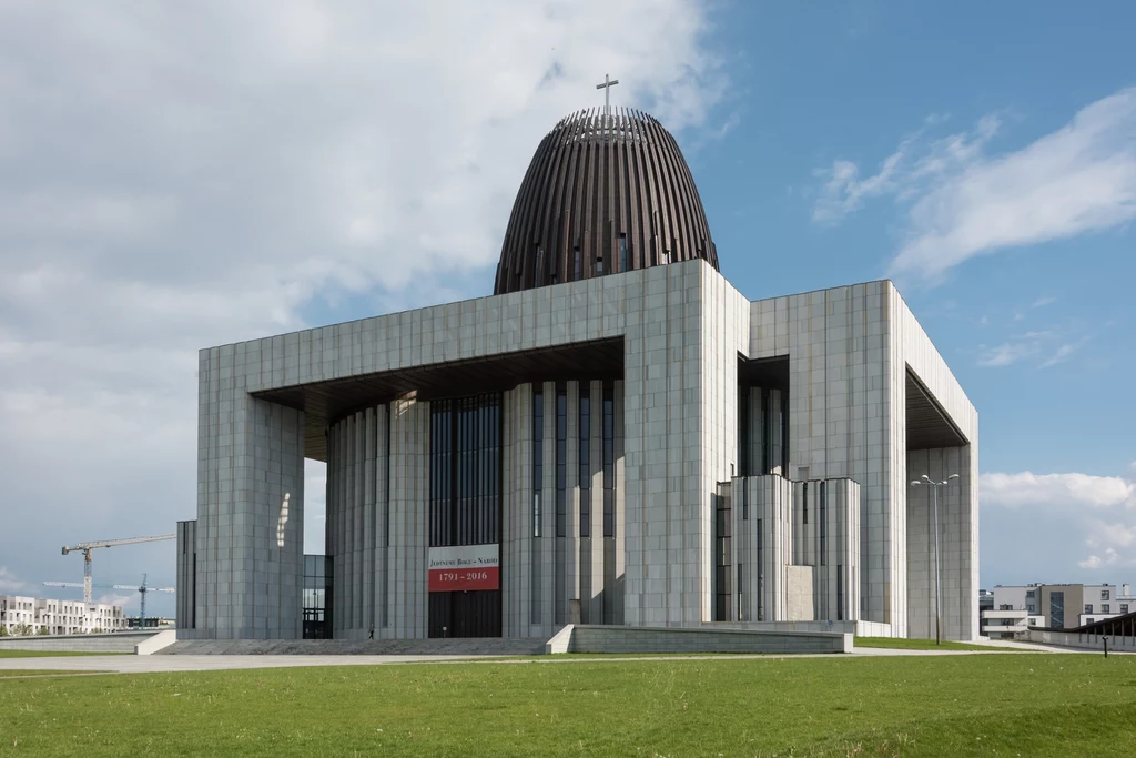 Świątynia Opatrzności Bożej, inaczej nazywana "Wyciskarką do cytryn" za sprawą wyjątkowego kształtu kopuły. Nowoczesny budynek o monumentalnej architekturze z wyraźnie widoczną dużą kopułą i krzyżem na szczycie, wykonany z jasnego kamienia, otoczony otwartą przestrzenią i zielonym trawnikiem, pod niebieskim niebem z lekkimi chmurami.