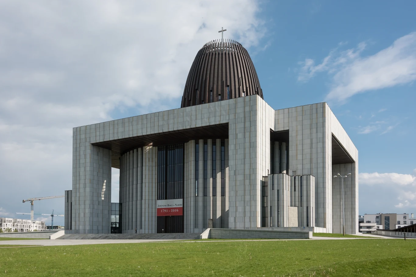 Nowoczesny budynek o monumentalnej architekturze z wyraźnie widoczną dużą kopułą i krzyżem na szczycie, wykonany z jasnego kamienia, otoczony otwartą przestrzenią i zielonym trawnikiem, pod niebieskim niebem z lekkimi chmurami.