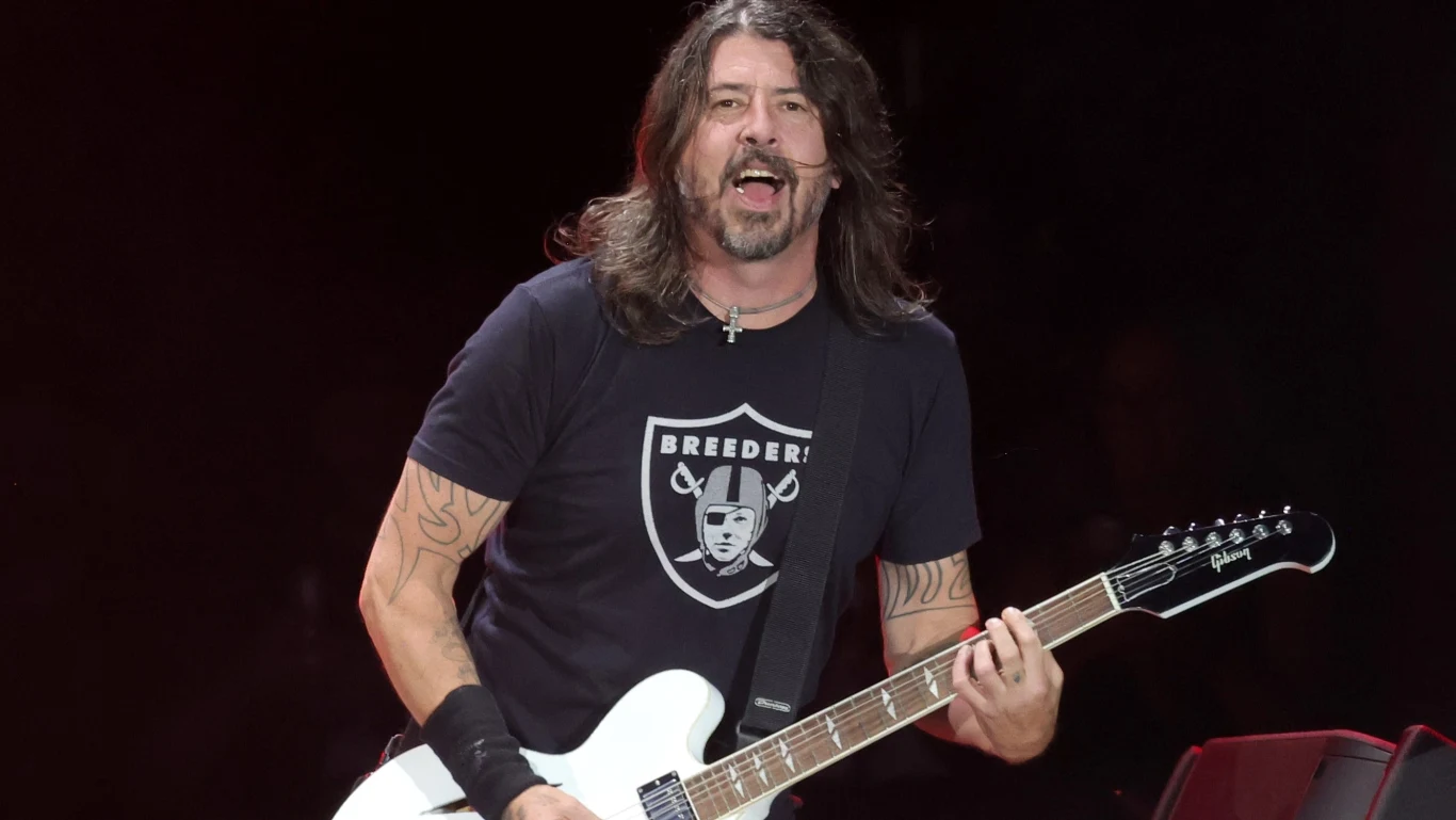 Foo Fighters powracają z nowym singlem! Mężczyzna o długich włosach gra na białej gitarze elektrycznej, ma na sobie czarną koszulkę z białym nadrukiem i tatuaże na ramionach, tło ciemne sceniczne.