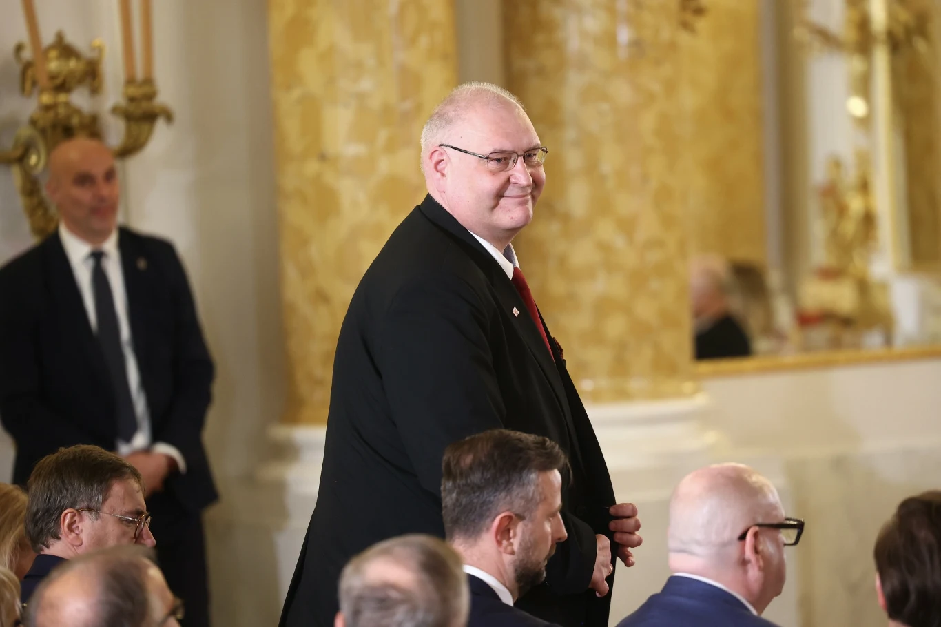 Oświadczenie majątkowe prezesa TK Bogdana Święczkowskiego Uśmiechnięty mężczyzna w garniturze idzie między siedzącymi ludźmi w eleganckim, złoconym wnętrzu, na drugim planie widać kolejnego mężczyznę przy ścianie.