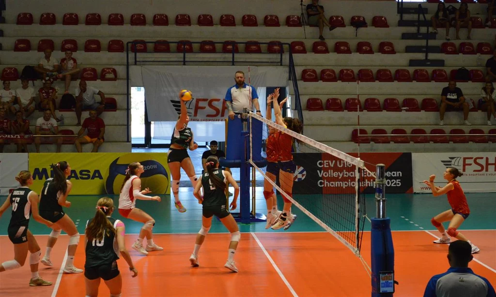 Drużyna siatkarek rozgrywa mecz na hali sportowej, jedna zawodniczka skacze do ataku, a inna próbuje zablokować piłkę przy siatce; zawodnicy w dynamicznym ruchu, na trybunach siedzi kilku widzów, sędzia obserwuje grę ze stanowiska ponad siatką.