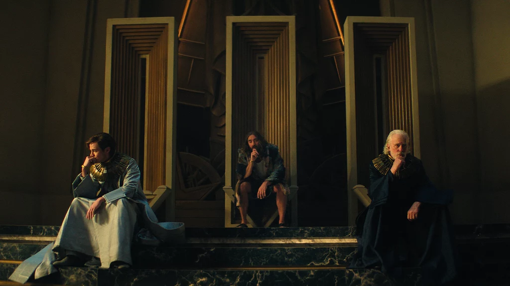 Cassian Bilton, Lee Pace i Terrence Mann w serialu "Fundacja". Trzech mężczyzn w ozdobnych szatach siedzi na wysokich, geometrycznie zdobionych tronach w monumentalnej sali, każdy z nich przyjmuje zamyśloną pozę, a całość charakteryzuje się atmosferą powagi i majestatu.