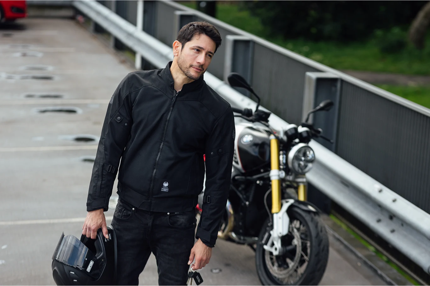 Mężczyzna w czarnej kurtce motocyklowej trzymający kask w ręku stoi na parkingu przy swoim motocyklu.