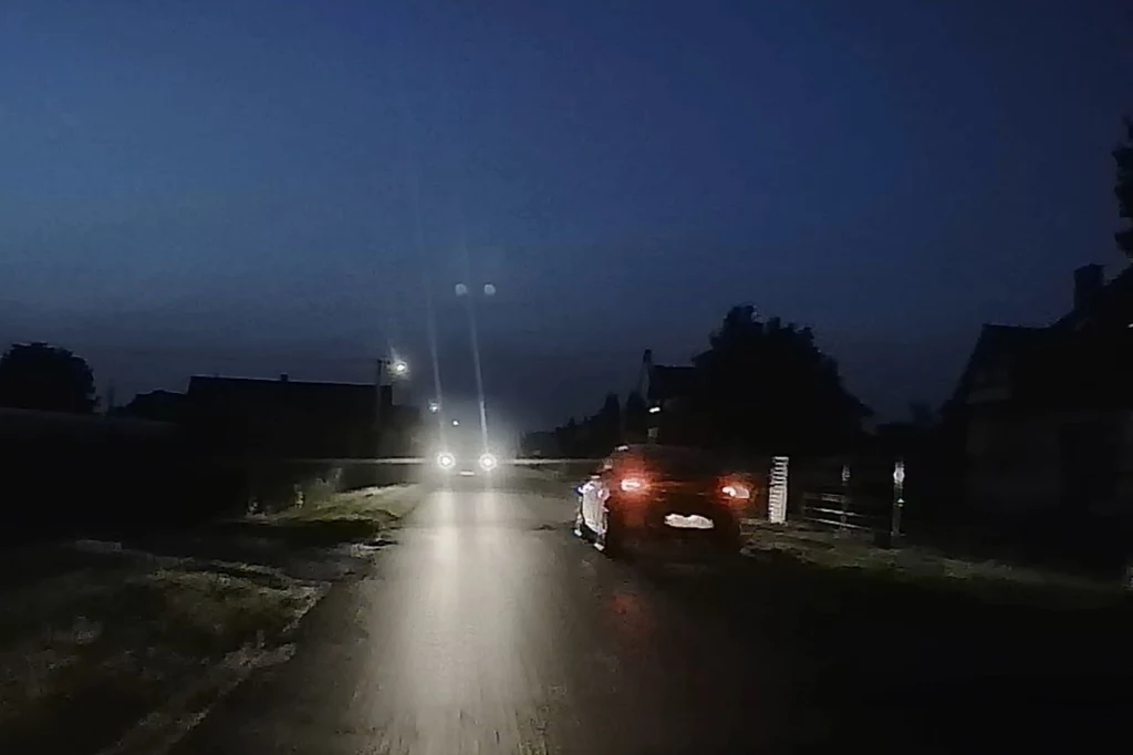 Jakość nagrań z tylnej kamery 70mai 4K Omni w nocy. Samochód poruszający się w nocy po słabo oświetlonej wiejskiej drodze, z reflektorami oświetlającymi asfalt, w tle widoczne światła kolejnego pojazdu oraz ciemne sylwetki domów i drzew.