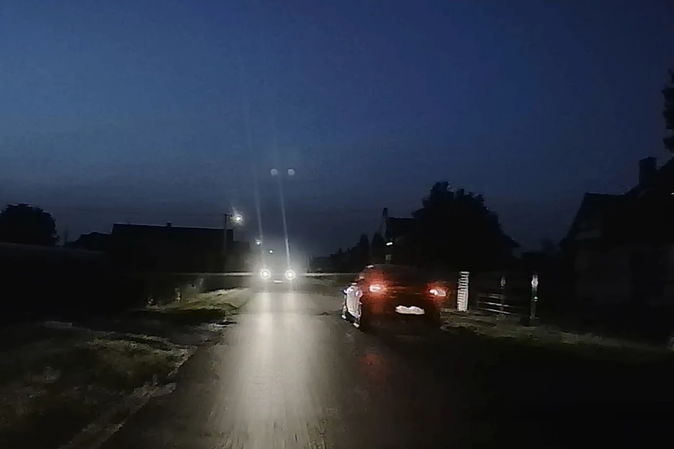 Samochód poruszający się w nocy po słabo oświetlonej wiejskiej drodze, z reflektorami oświetlającymi asfalt, w tle widoczne światła kolejnego pojazdu oraz ciemne sylwetki domów i drzew.